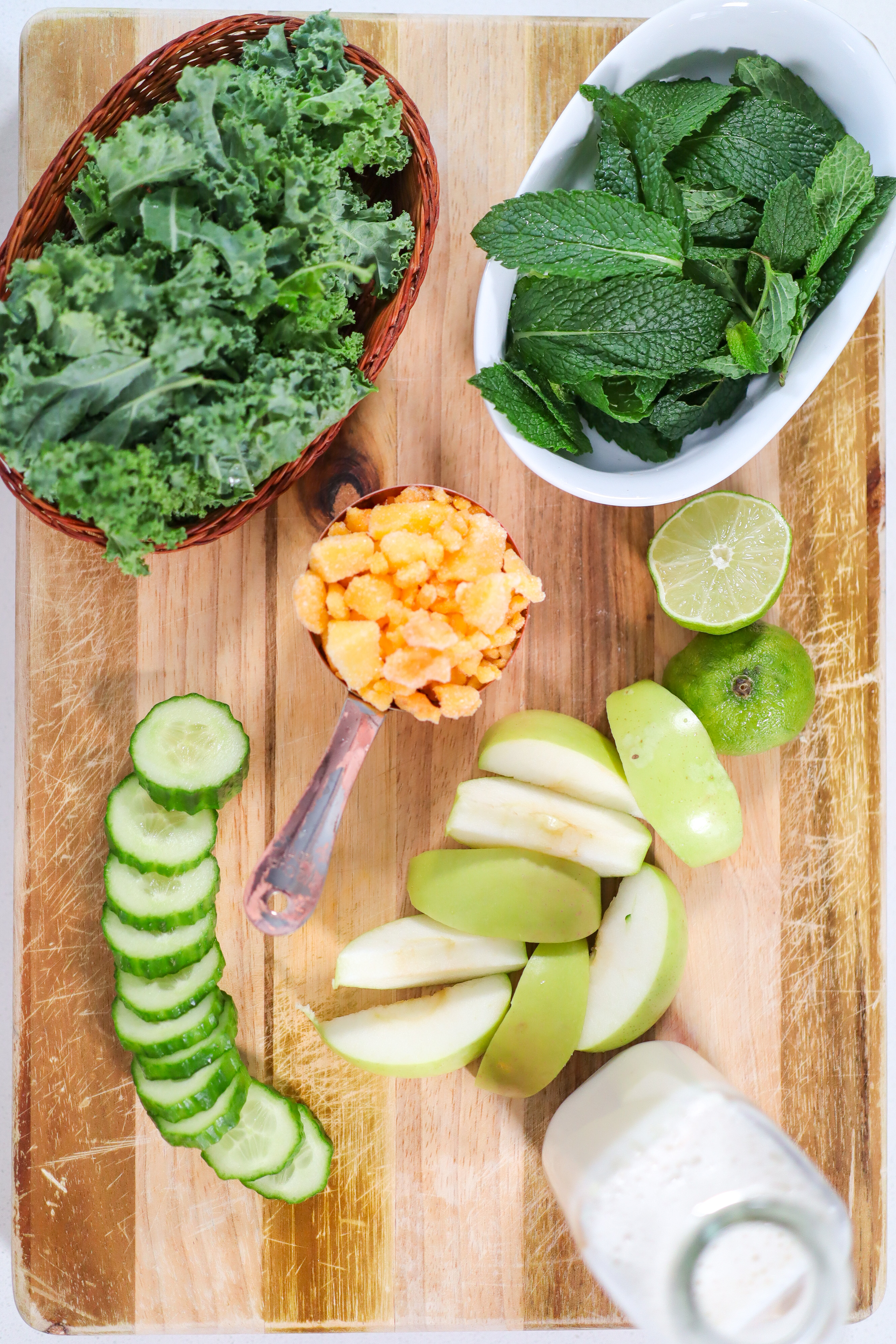 wood cutting board with kale, mint, cucumber, apple, milk, and mango on it