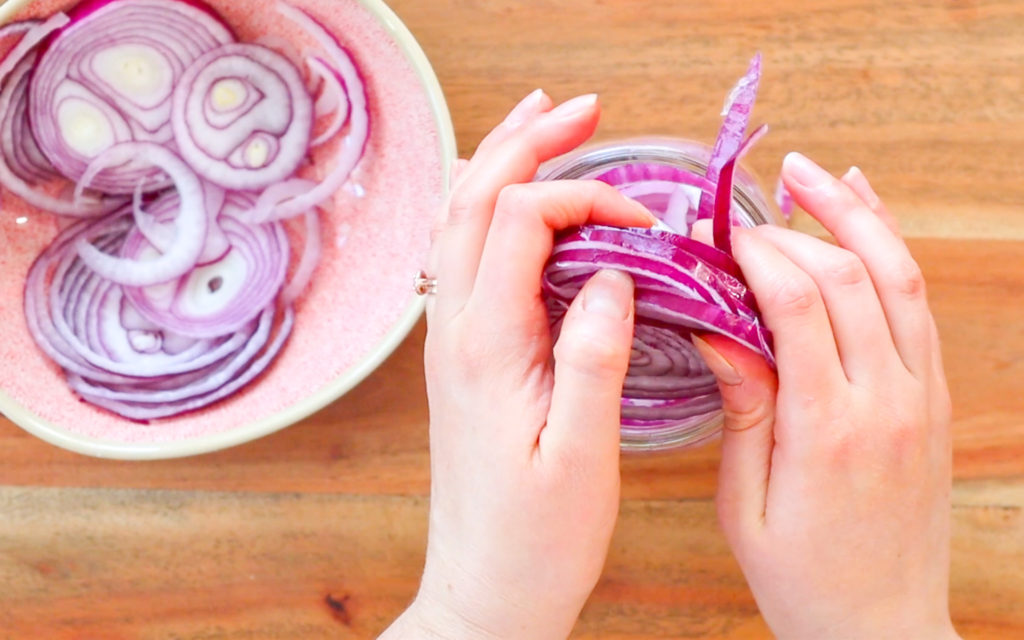 hands stuffing sliced red onions into a glass jar