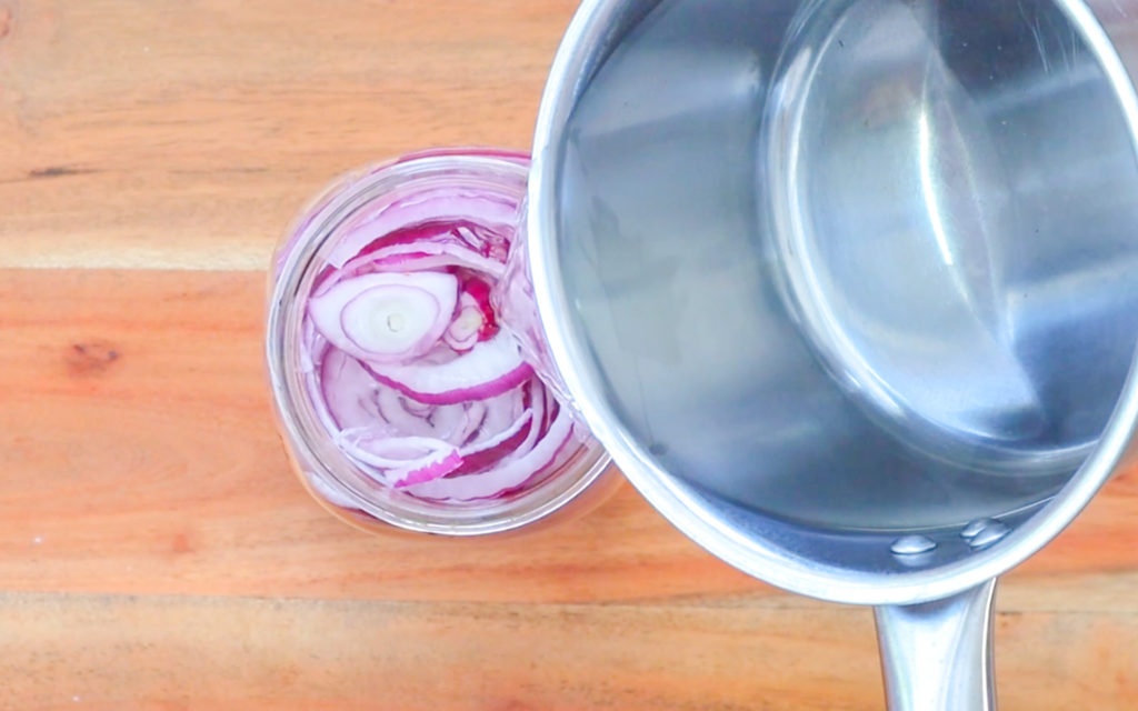 A small saucepan pouring liquid into a jar of sliced red onions