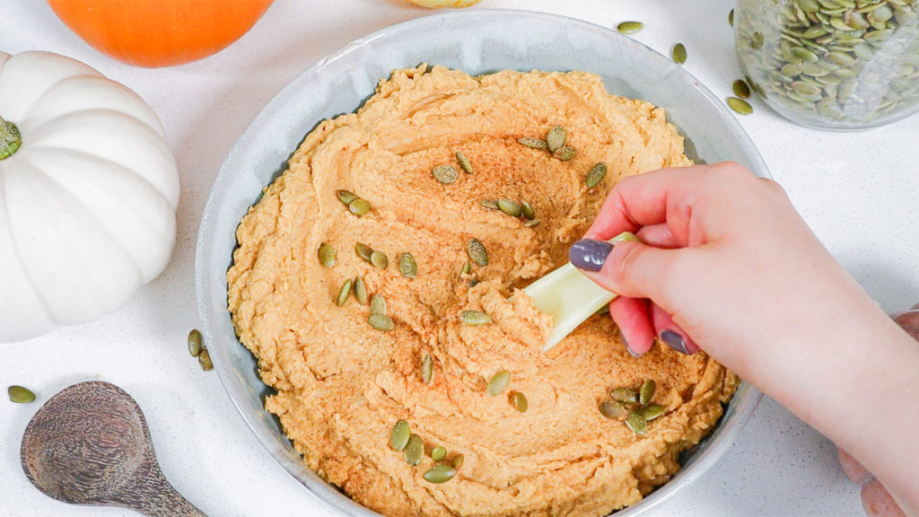 Hand holding celery stick scooping up some pumpkin spice hummus.