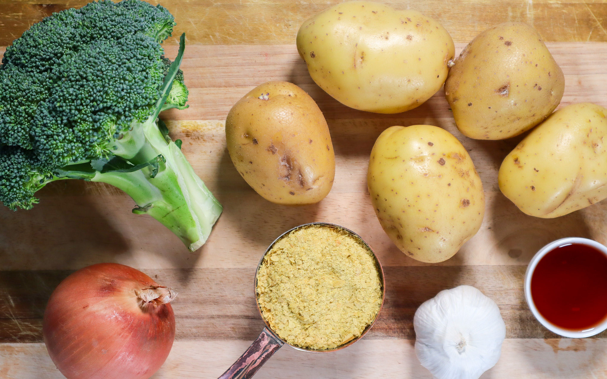 whole yellow potaotes, broccoli, an onion, garlic, red wine vinegar and garlic on a wood cutting block.